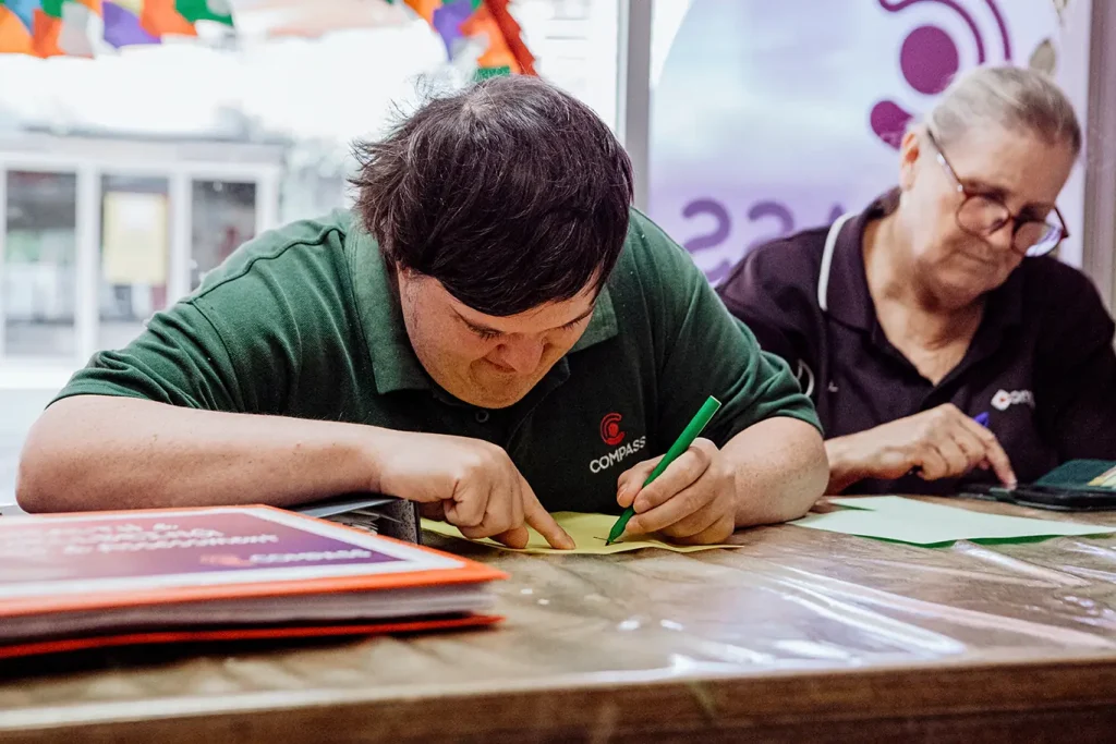 A person with Down’s syndrome participates in skills-based training, writing with a green marker at a table while an older woman beside them also writes. Colourful decorations hang in the background.