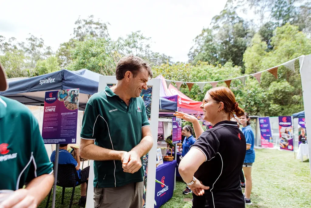 Two people stand and talk at an outdoor event with informational stalls and tents, including one for Disability Services. Others mingle in the background. Trees and bunting decorate the area, creating a festive, community atmosphere.