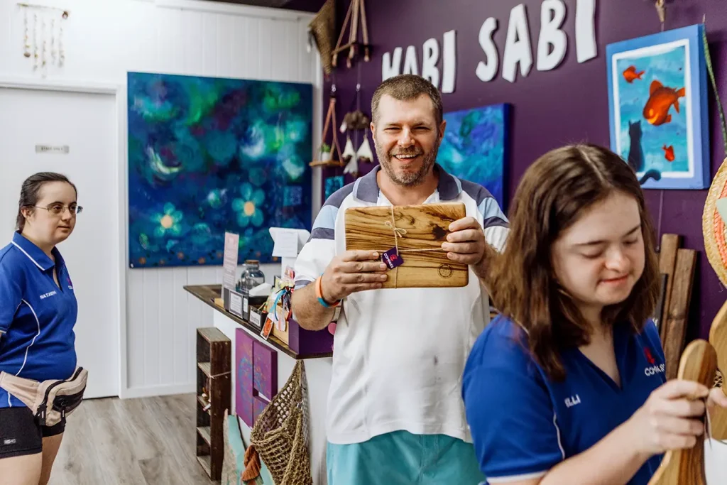 Three people smile and hold crafted wooden boards in a colourful art studio with purple walls, paintings, and the words WABI SABI displayed in the background, embodying a "What If" spirit of creativity and inclusion.