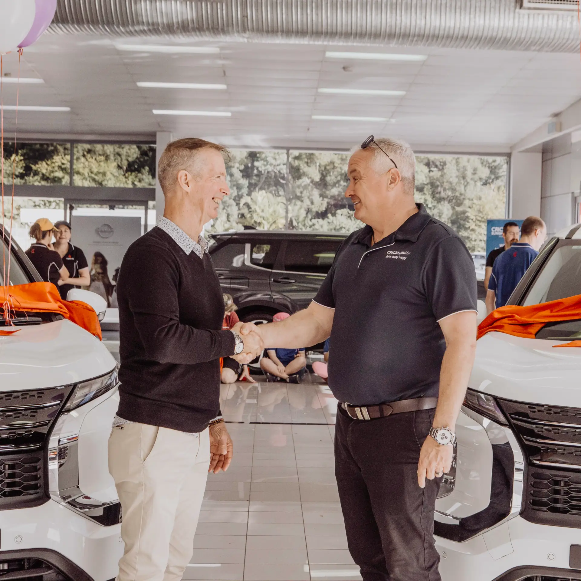 Two men are shaking hands and smiling at each other inside a car showroom, possibly celebrating the success of a campaign, as they stand between two white cars with orange ribbons on the bonnets. Other people and vehicles are visible in the background.