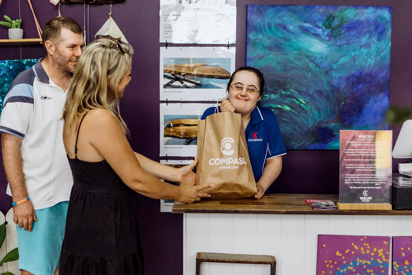 A smiling young woman in a blue Compass uniform hands a paper bag to a blonde woman across the counter, with a man nearby. Colourful artwork decorates the purple wall, reflecting the creativity of the What if Campaign.